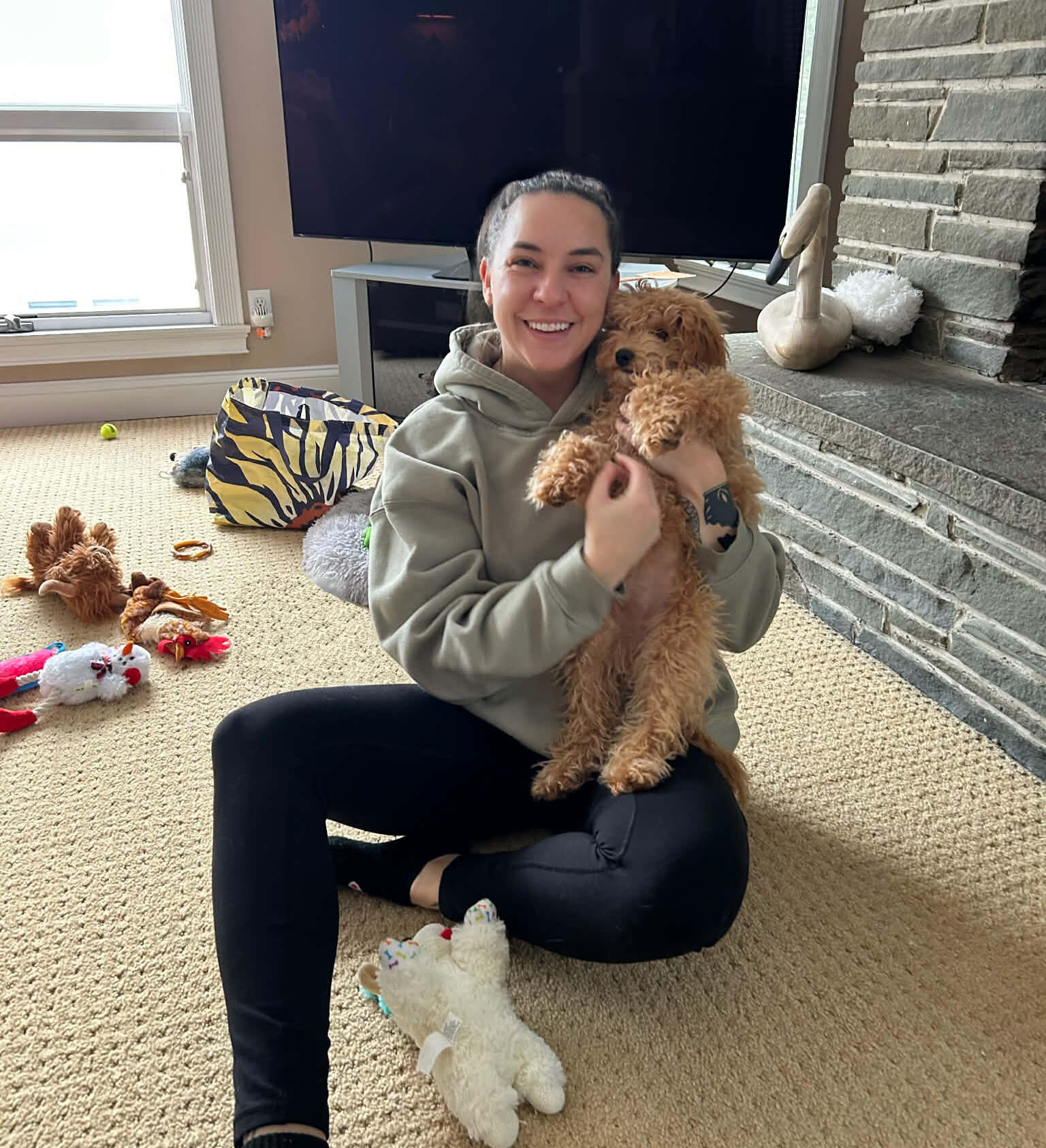 Sophie Williams enjoying some puppy playtime with Ruby. Sophie Williams smiling, sitting cross-legged on a carpeted living room floor, holding a small fluffy brown puppy, with dog toys scattered around and a stone fireplace in the background.